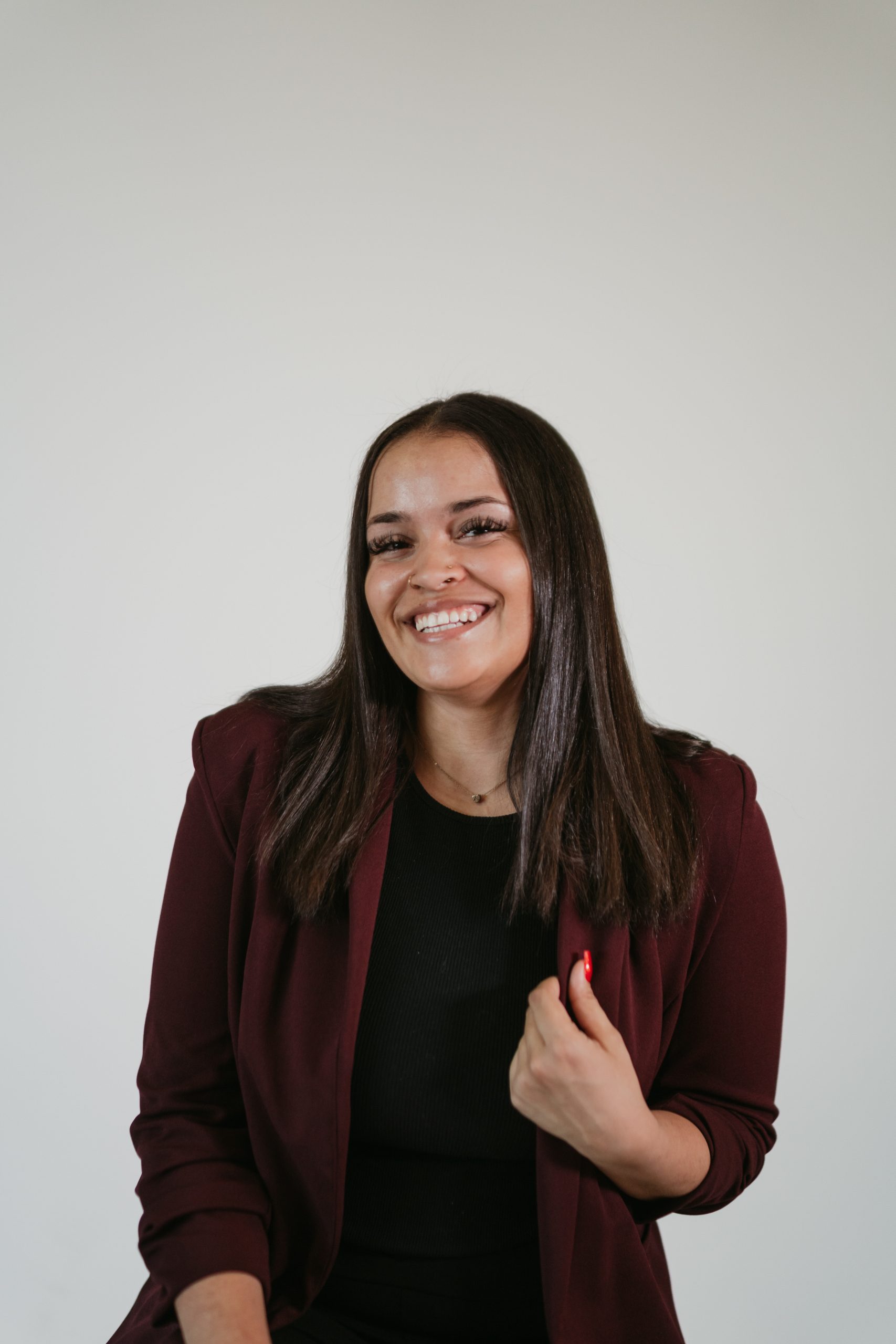 Paris Baptiste sitting with a dark top and blazer with long dark hair and smiling